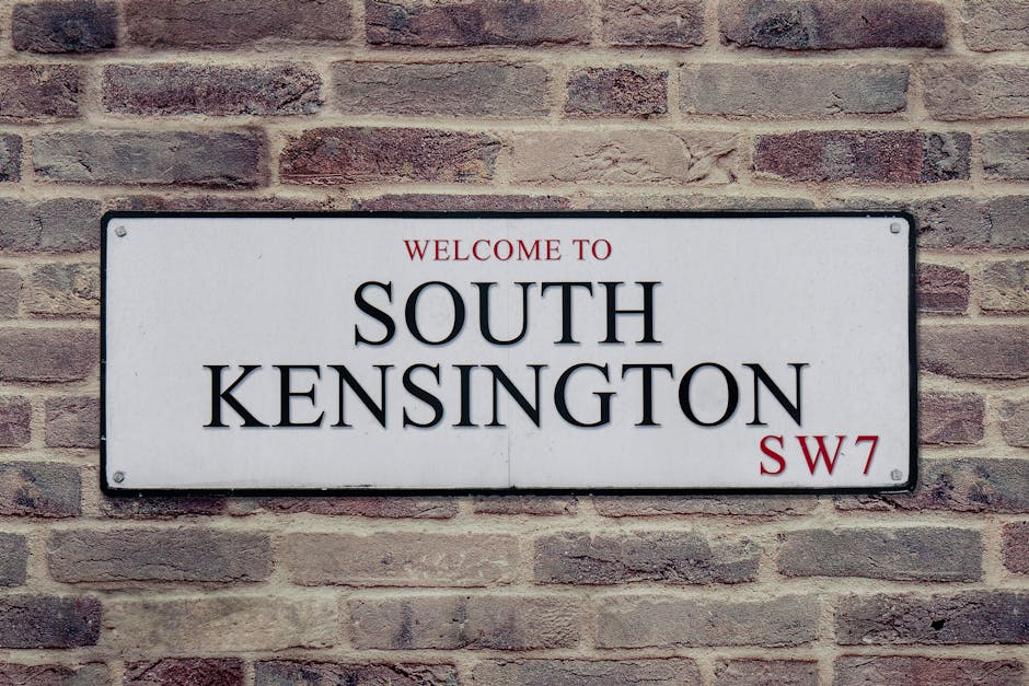 A rectangular metal street sign mounted on a red brick wall, displaying the text 'WELCOME TO SOUTH KENSINGTON SW7' with 'WELCOME TO' in red uppercase letters at the top, 'SOUTH KENSINGTON' in large bl